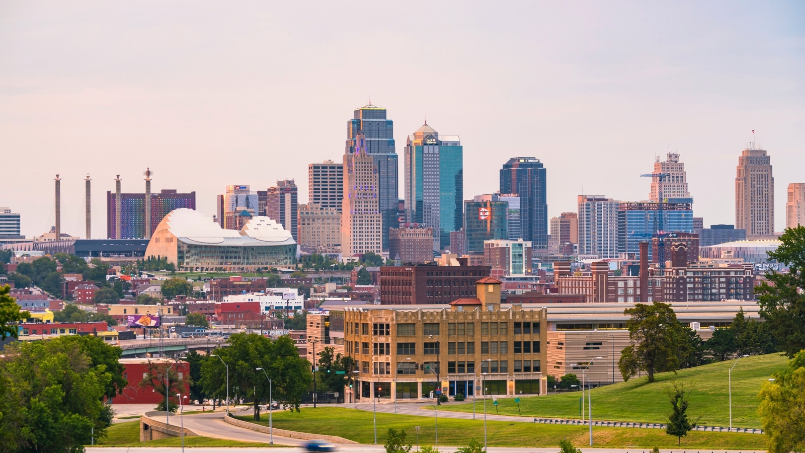 The Kansas City skyline at sunset features a blend of modern and historic buildings. The cityscape includes tall skyscrapers with reflective glass exteriors and shorter, older structures with brick facades. The sunset casts a warm, golden hue over the buildings, enhancing the visual appeal.