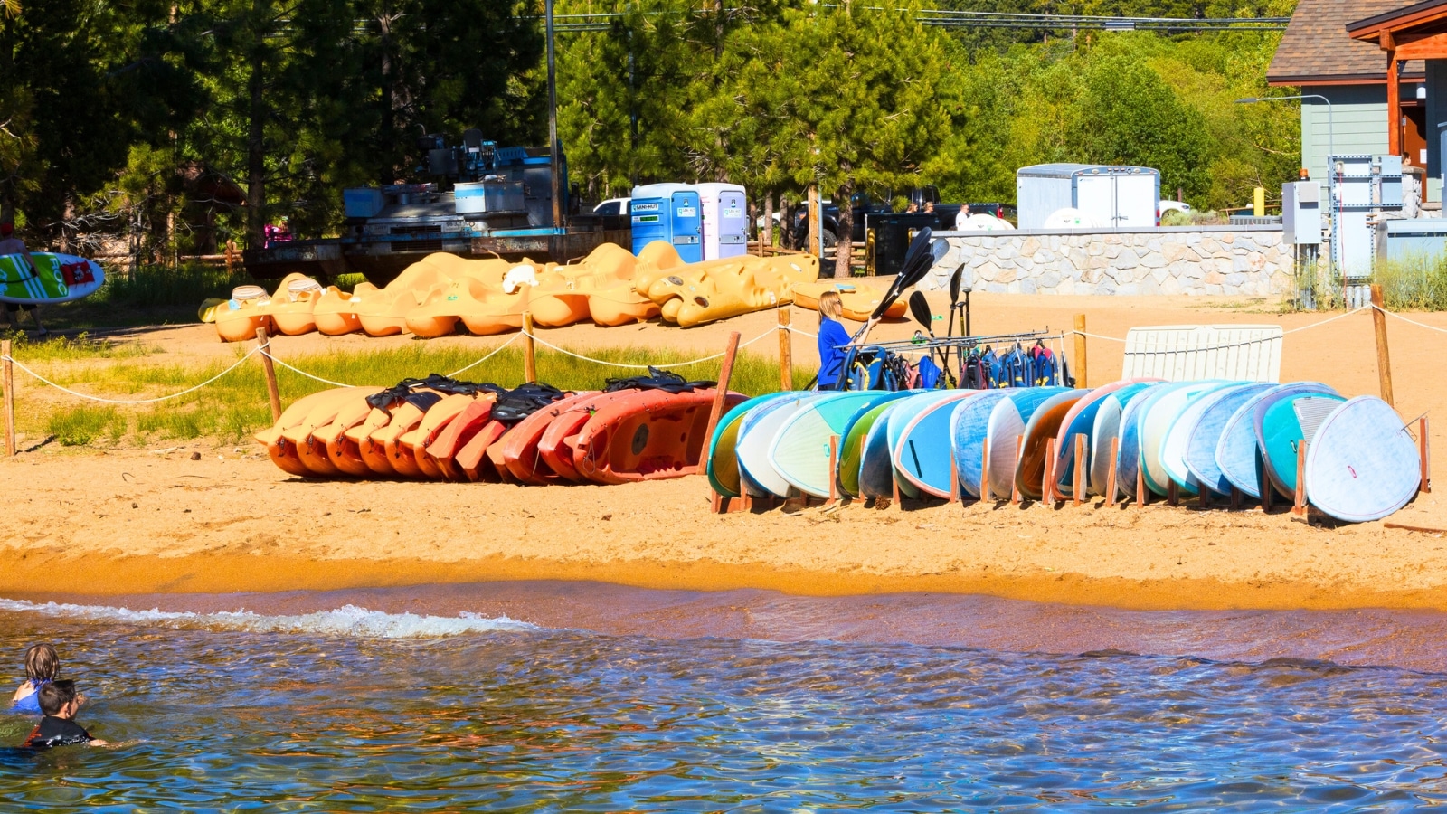 The image depicts a scene at Lake Tahoe in Nevada, USA, showcasing a stack of kayaks available for rent on the beach.