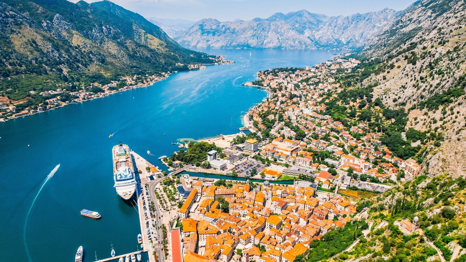 Bay of Kotor in Montenegro. In the center of the bay is a tranquil expanse of water, surrounded by large hills on both sides. To the right, a town with brown-roofed buildings adds a touch of traditional charm to the landscape. At the pier, a ship and several boats are moored.