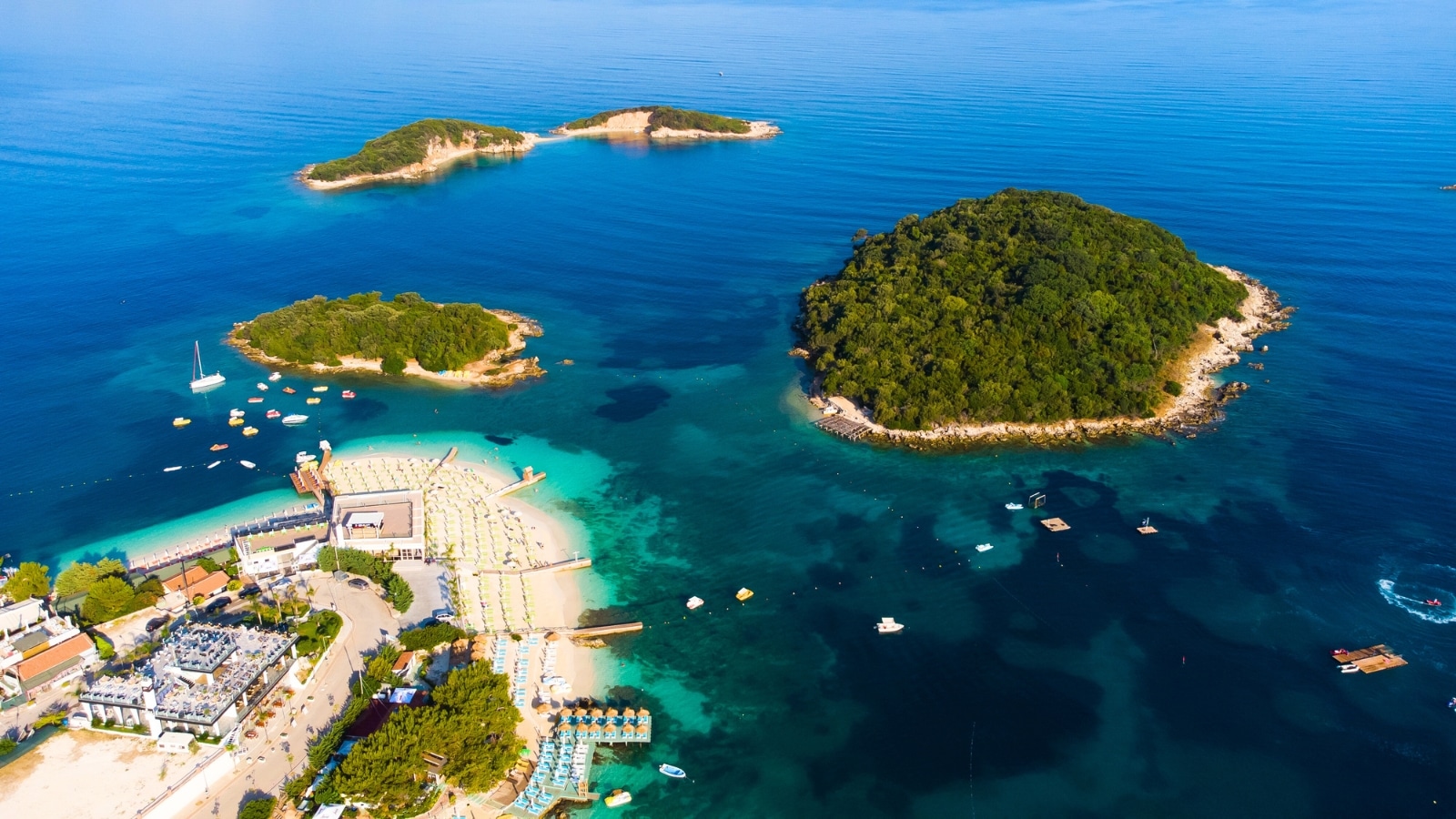 The drone's view reveals Ksamil Beach in Albania, featuring several islands adorned with lush greenery scattered across the sea. Numerous boats dot the azure waters. The beach itself boasts golden sands contrasting with the deep blue sea.