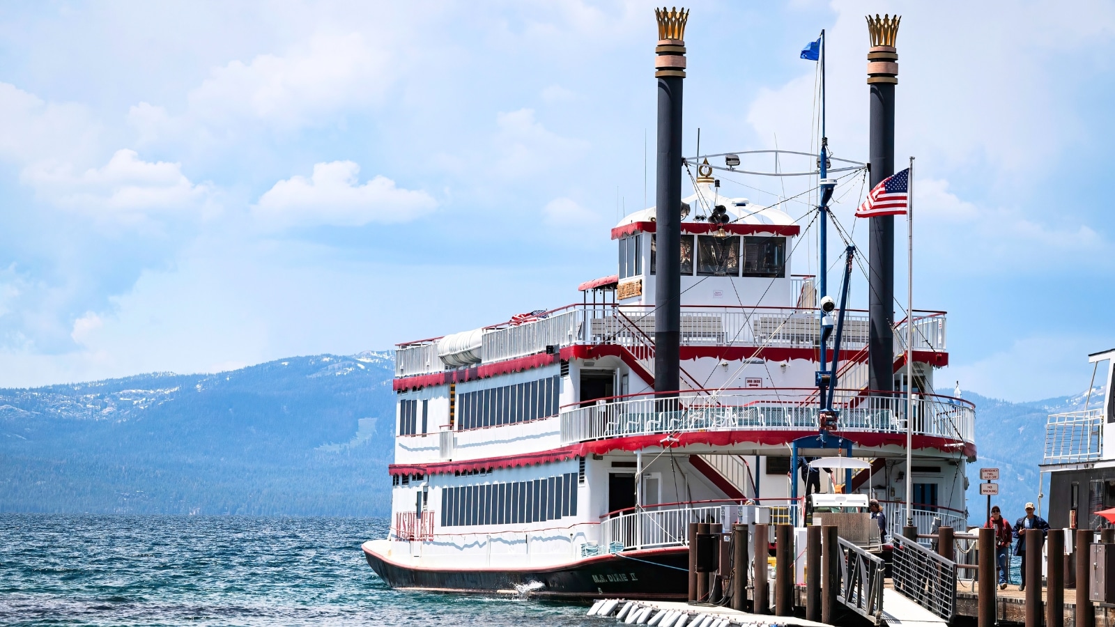 Lake Tahoe with the M.S. Dixie II, a large ship, prominently featured. The ship is predominantly white with red stripes, and the USA flag is visible. Surrounding the ship are the serene waters of Lake Tahoe, reflecting the blue sky above.