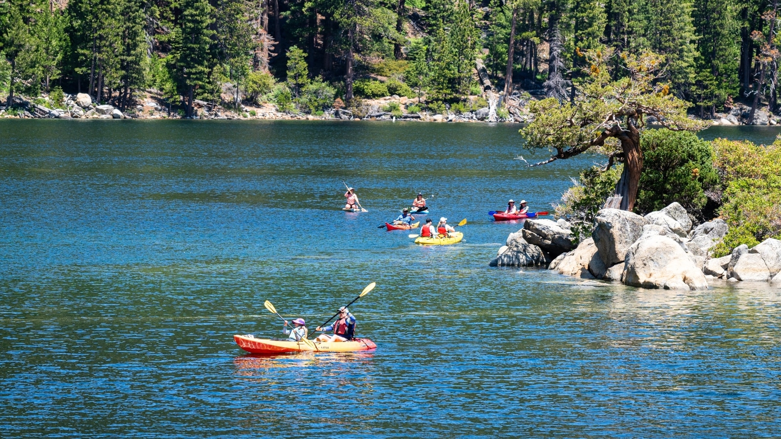 The image depicts people kayaking on South Lake Tahoe in California, USA. Six kayaks, colored in red and yellow, are visible in the frame as they navigate the serene waters. Tall pine trees and other foliage form the backdrop, highlighting the natural beauty of the lake's surroundings.