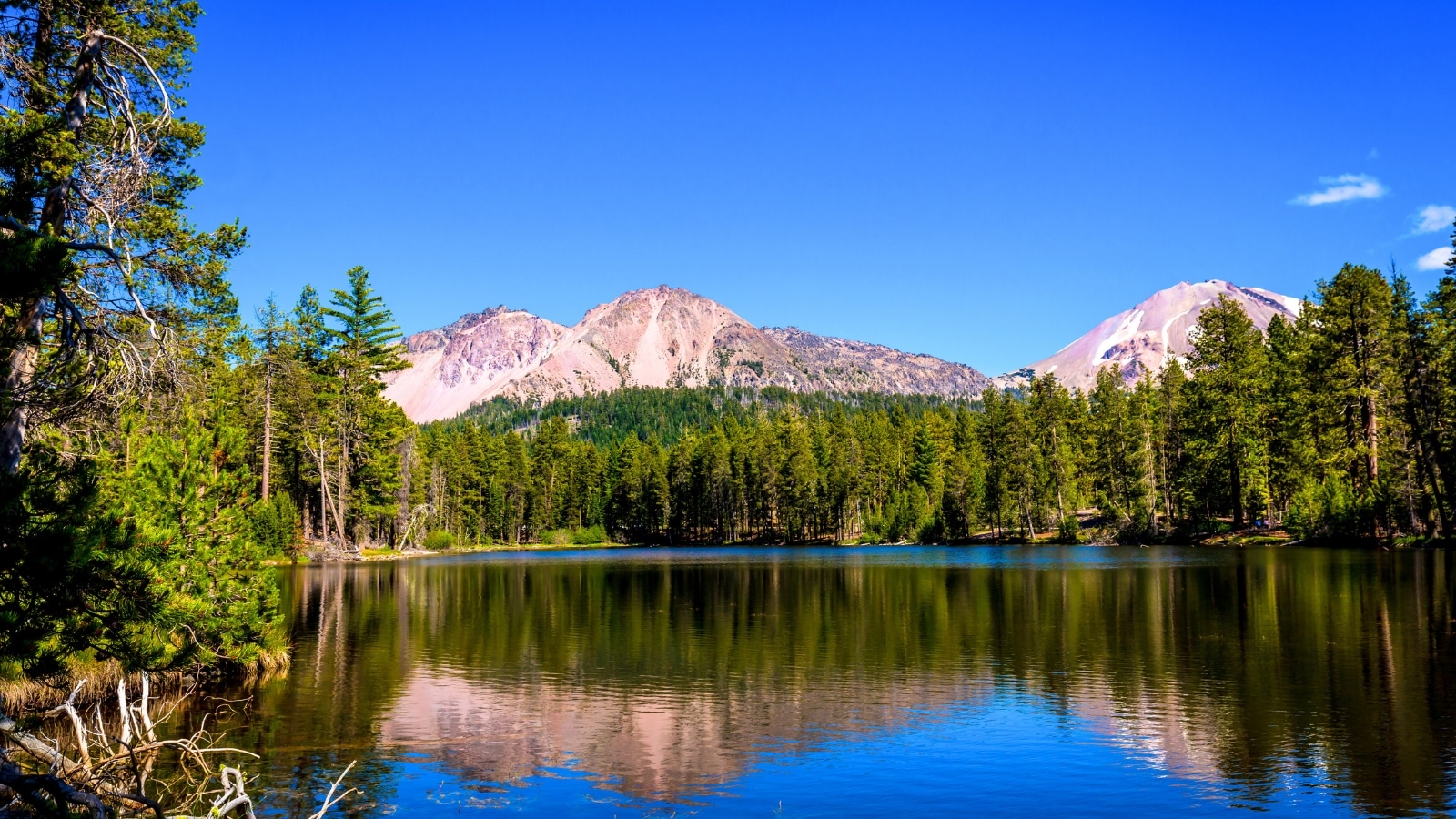 Chaos Crags and Lassen Peak reflected in Reflection Lake, situated in Lassen Volcanic National Park, California. In the foreground, the lake's calm waters mirror the rugged beauty of the volcanic peaks. Beyond the lake, a dense forest provides a natural backdrop to the scene. Further in the distance, rocky mountains rise, completing the panoramic view of the park's diverse terrain.
