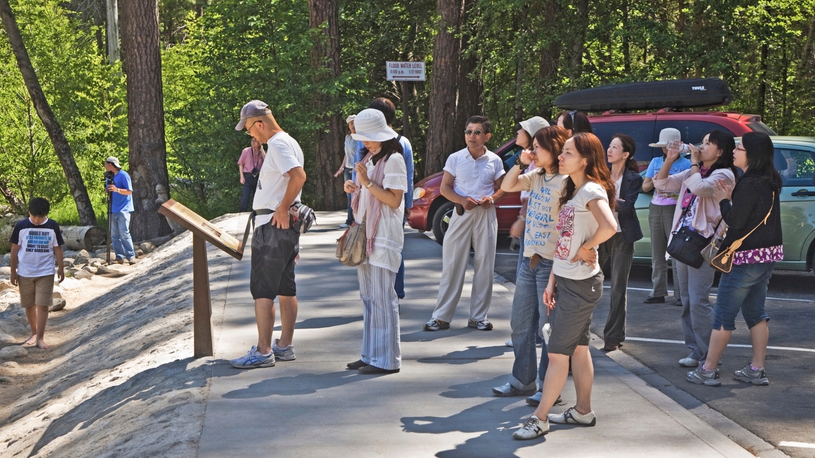 Yosemite National Park in the United States. In the foreground, numerous people are visible near a parking lot, engaging in various activities. Some are observing the park's signage, while others are gazing around the park. One individual is seen capturing photographs. 
