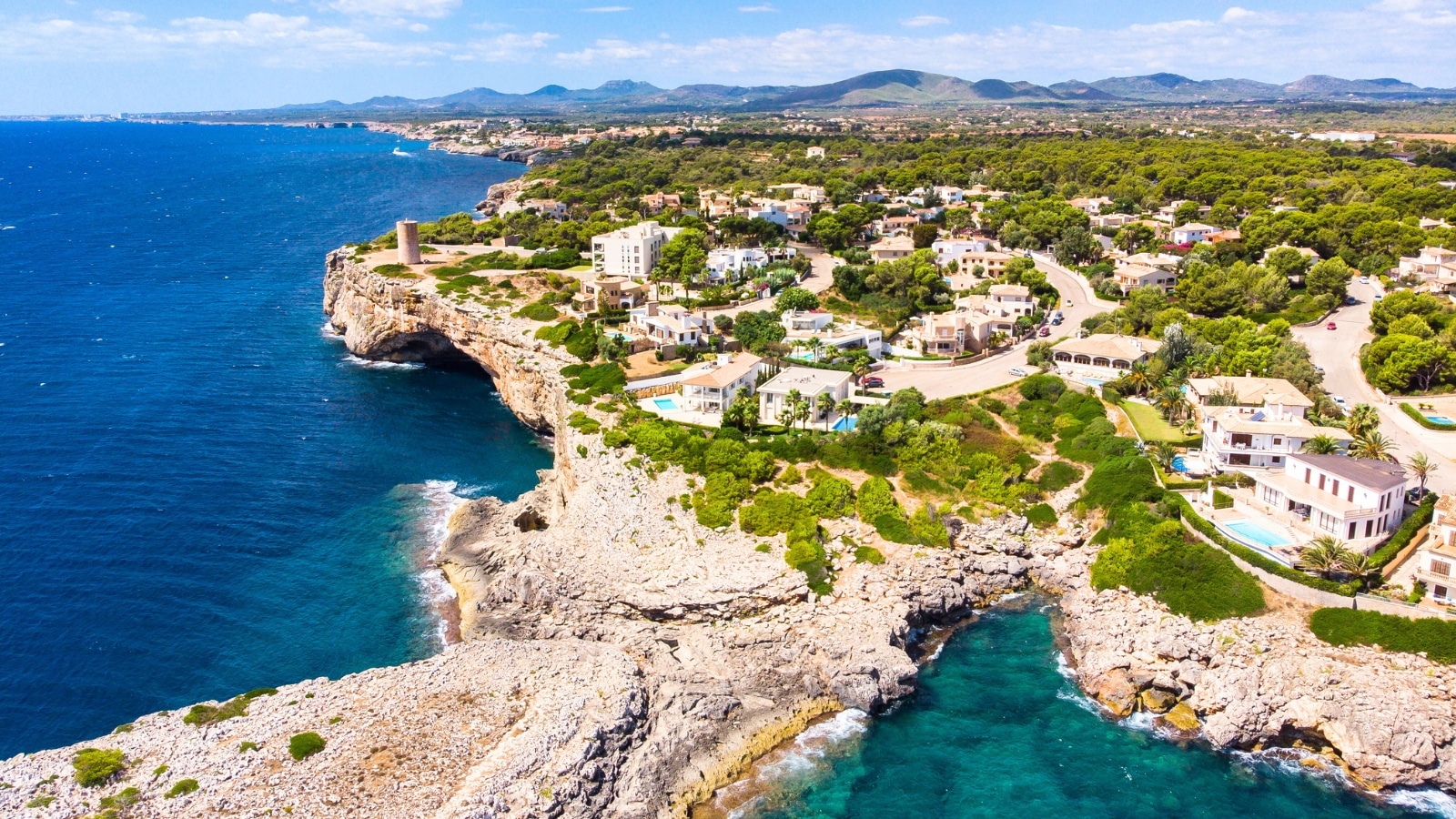 An aerial view reveals Porto Cristo, a coastal area on Mallorca, part of the Balearic Islands in Spain. The scene includes villas lining the coast and a natural harbor. The villas exhibit a variety of architectural styles, with white facades and terracotta roofs predominating.