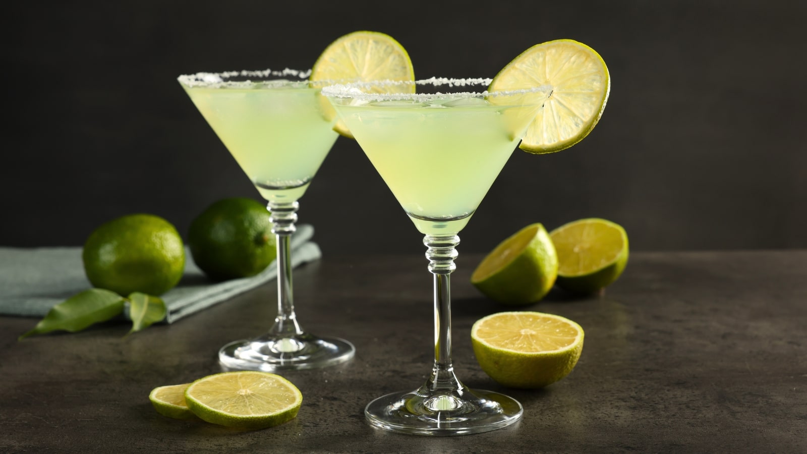 On the table, there are two glasses filled with margarita cocktails, accompanied by several limes in the background. The cocktails appear refreshing, with a vibrant green hue from the lime and a hint of golden color from the drink itself.