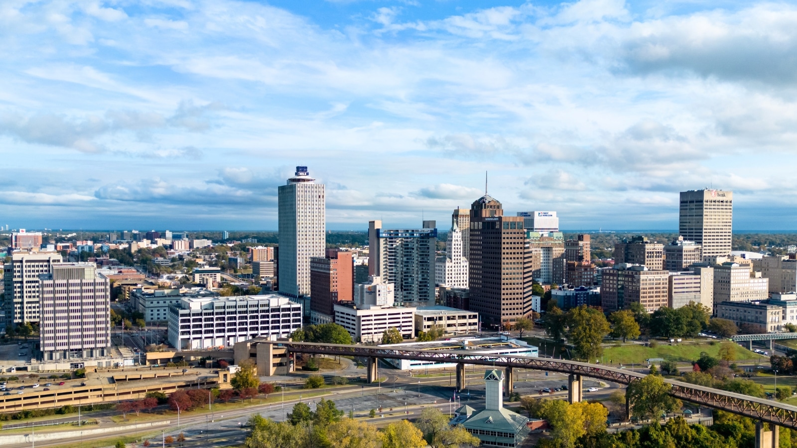 The panoramic view over Memphis, Tennessee, features a blend of urban and natural elements. The cityscape includes modern skyscrapers and historic buildings with varying architectural styles. The Mississippi River flows alongside the city.