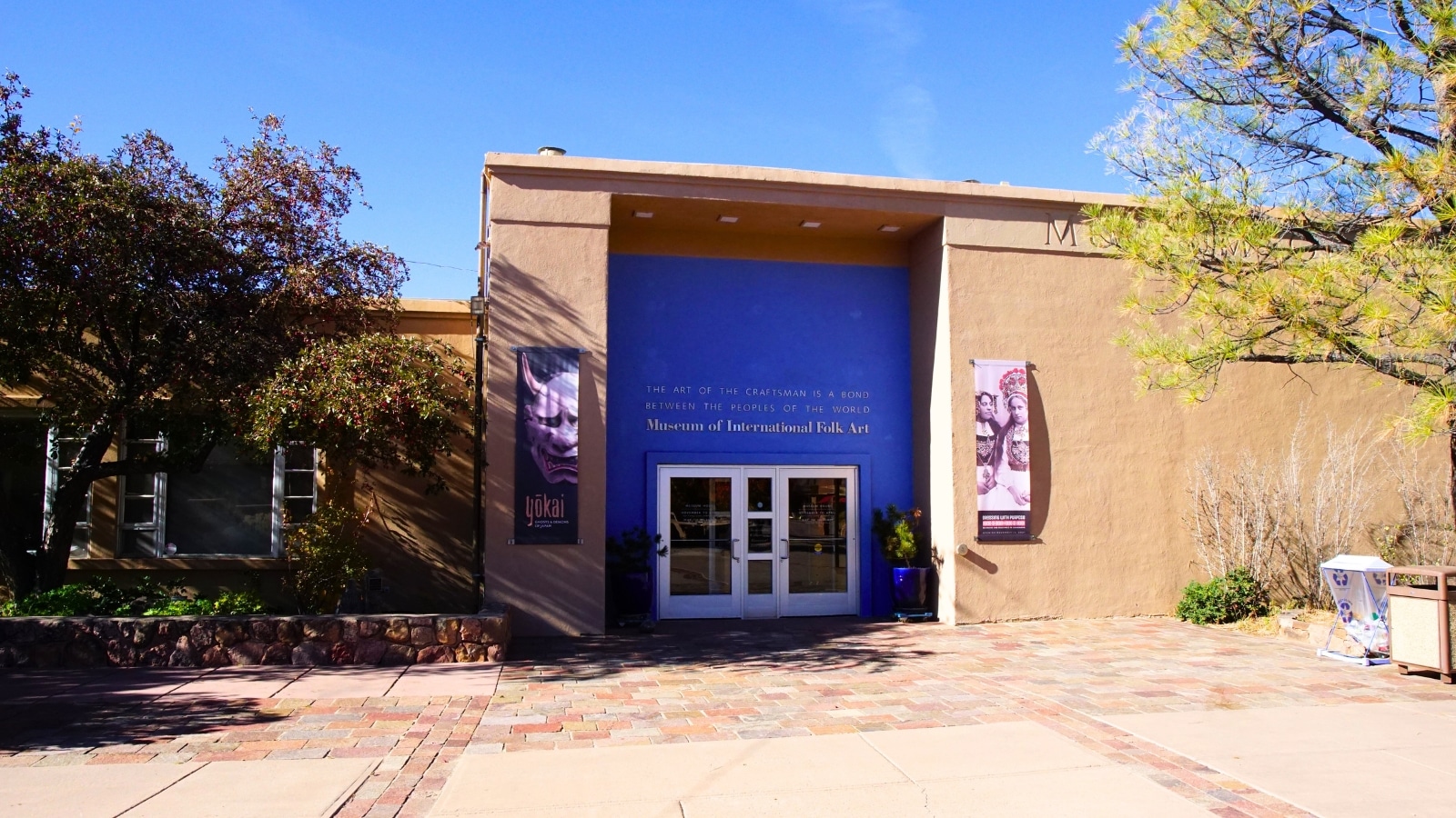 The entrance to the Museum of International Folk Art in Santa Fe, New Mexico, features a distinctive facade with a blend of colors. The foreground showcases a blue section of the facade, while the front door, adorned with glass panels, is in white. Surrounding the entrance, the rest of the building's facade is in a beige hue, creating a harmonious yet striking visual contrast.