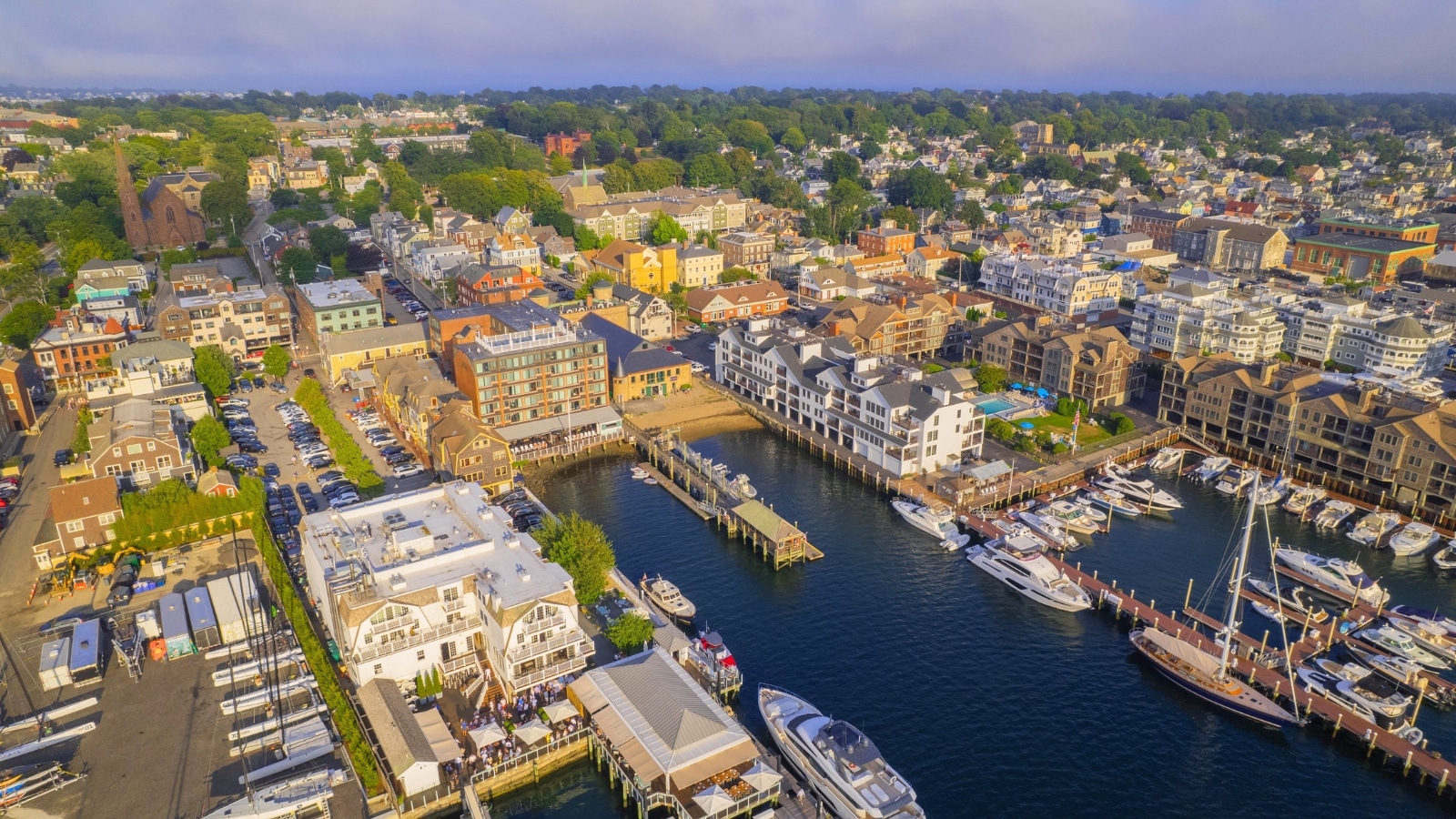 An aerial shot of Newport Harbor in Rhode Island reveals docked boats and a landscape. The city is surrounded by abundant greenery and trees, with the city port prominently featured in the foreground. The port is bustling with boats and ships.