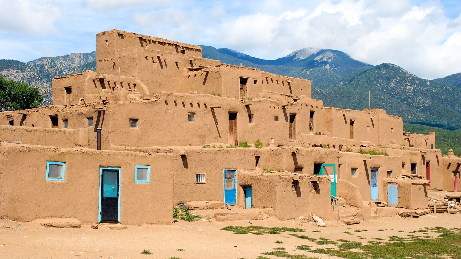 The image depicts an Old Pueblo structure set against a backdrop of rugged mountains and cloudy skies. The structure features traditional adobe architecture, with thick walls and a flat roof, typical of the region's historical buildings.
