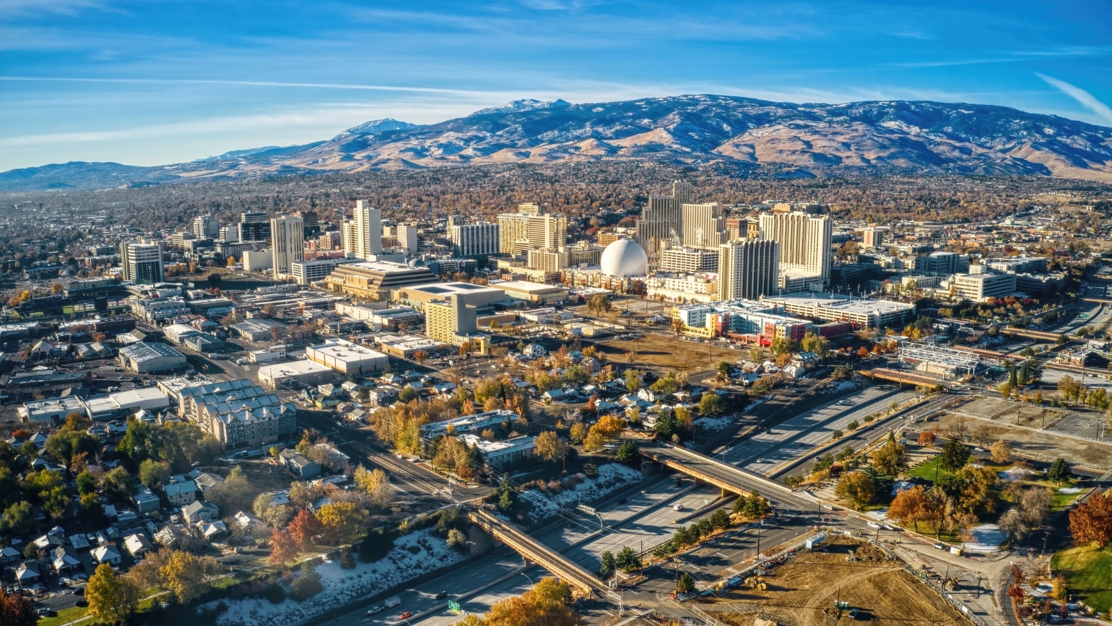 Reno, Nevada, offers a panoramic view from a drone, highlighting the city's blend of urban and natural elements. The cityscape features a mix of buildings, roads, and green spaces, indicating a balance of development and vegetation. The backdrop includes mountains, presenting a stark contrast with the city's structures.