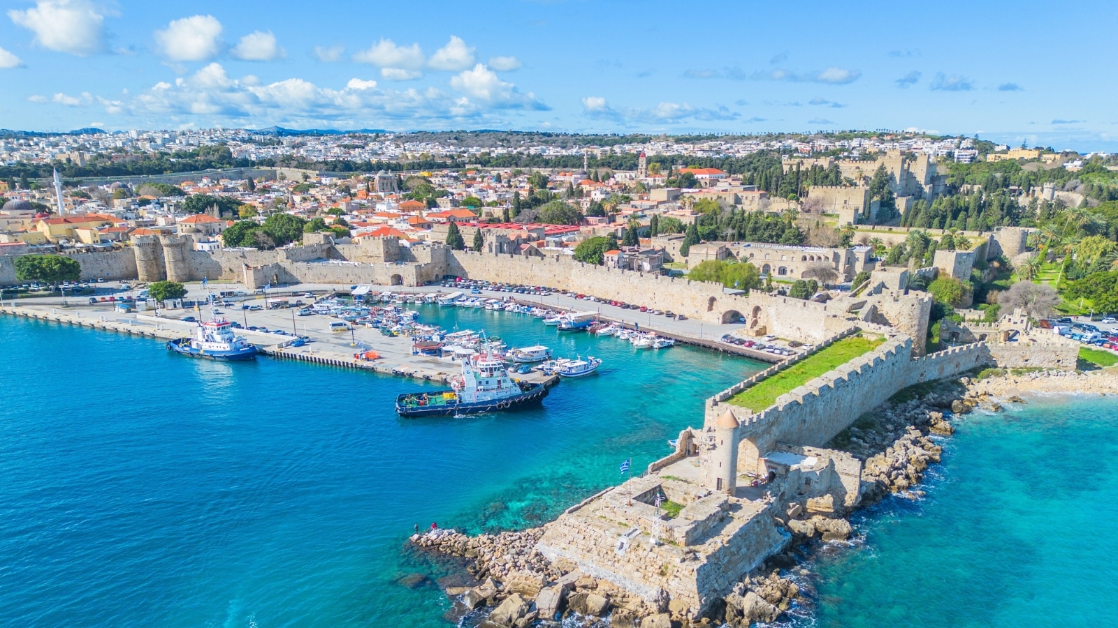 The image is an aerial view captured by a drone, showcasing the marina of Rhodes Island in Greece. The scene features numerous boats and yachts docked in the port, creating a vibrant maritime atmosphere.