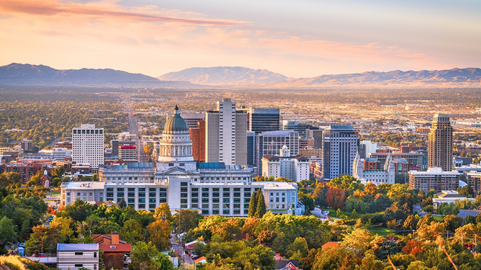 Salt Lake City, Utah, USA, displays a downtown city skyline at dawn. The cityscape features a mix of modern and historic buildings, illuminated by the soft light of early morning. The backdrop consists of mountains and hills.