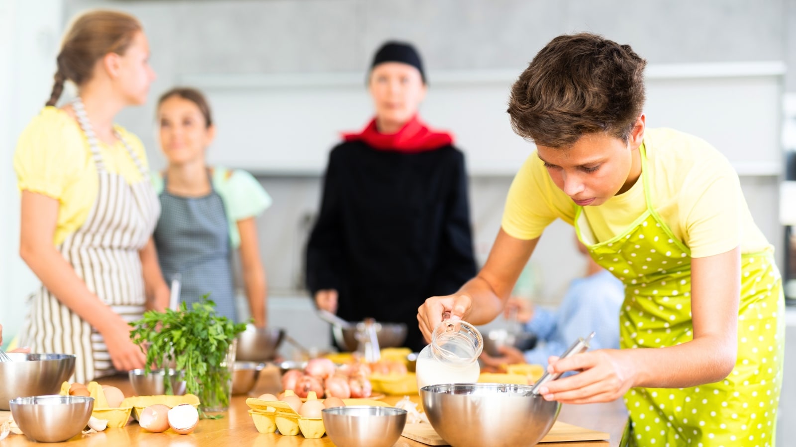 The image depicts a cooking lesson led by a chef in a black suit, surrounded by attentive students. On the table, there are eggs and bowls.