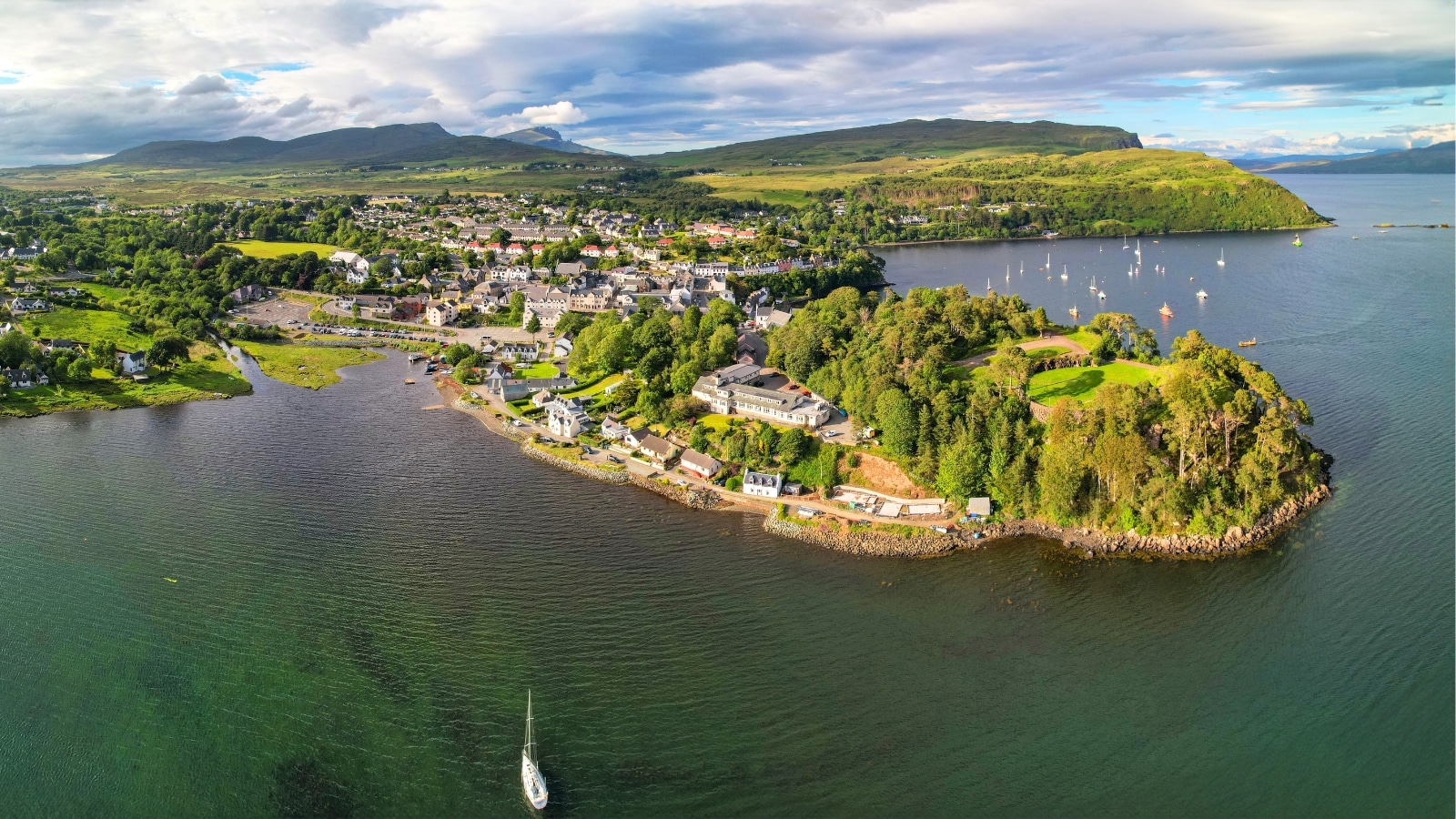 The image captures a peaceful bay near the picturesque island town of Portree. The island is adorned with lush greenery, including numerous trees that dot its landscape. In the background, small hills rise gently, adding depth to the scenic view. The foreground showcases the calm sea.
