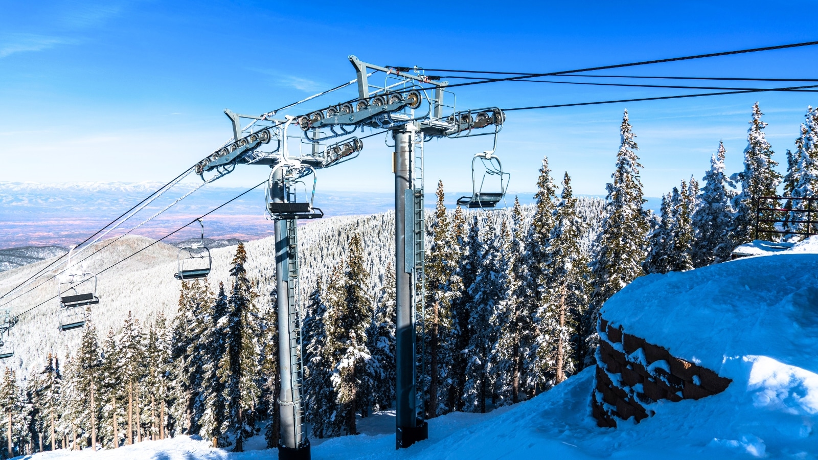 Atop the snow-covered Santa Fe Mountains stands a ski lift, its iron structure contrasting against the clear skies. The scene is adorned with a blanket of pristine snow, dotted with tall fir trees that add a touch of green to the wintry landscape.