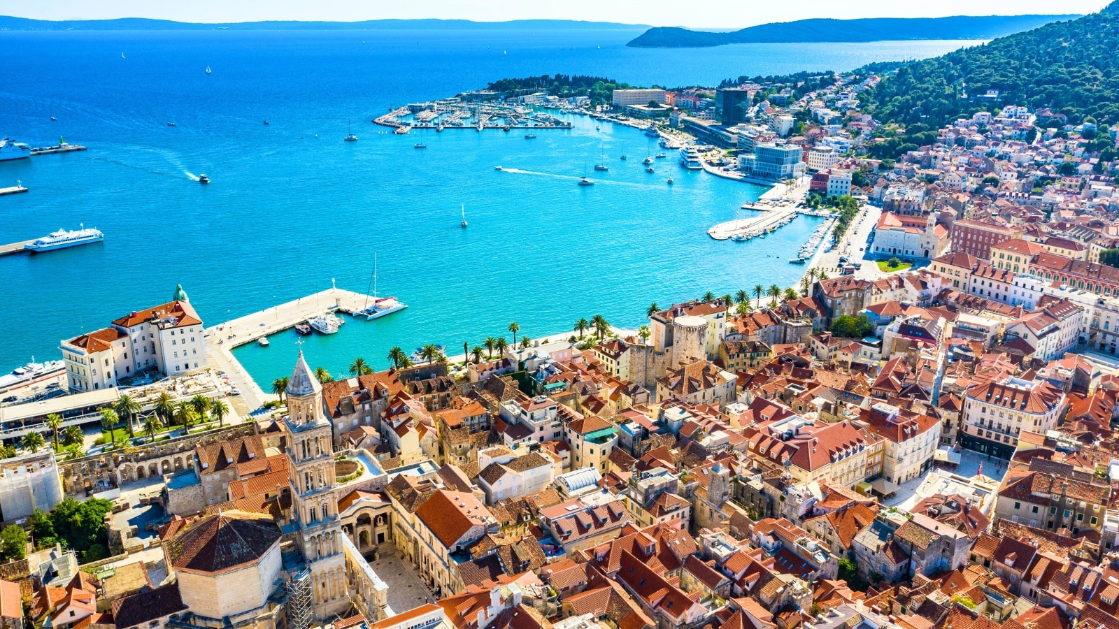 The panoramic view from a drone captures Split, a town in Dalmatia, Croatia. The scene includes a mix of historical and modern buildings, with red-tiled roofs and stone facades typical of Mediterranean architecture. The town is situated along the coastline, with the Adriatic Sea visible in the background. The waters showcase varying shades of blue, from deep azure to lighter turquoise.