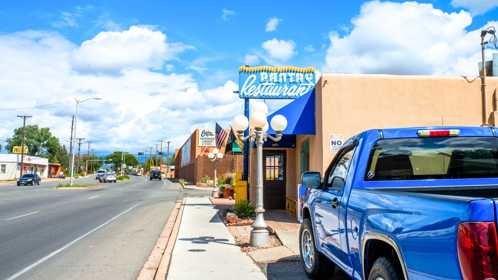 The Pantry restaurant in Santa Fe, New Mexico, is highlighted by a prominent blue sign, standing out against the backdrop of the cityscape. In the foreground, a portion of a blue car adds to the color scheme of the scene. The street, visible in the distance, adds a sense of urban ambiance to the setting.