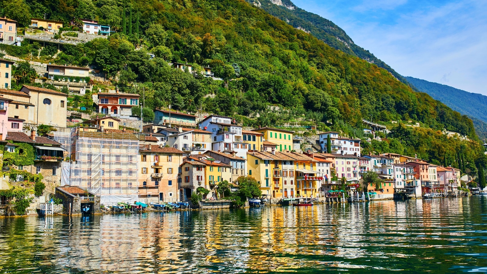 The scenic view of Gandria village near Lugano, in the canton of Ticino, Switzerland, offers a picturesque scene from the lake. The village consists of charming old buildings with terracotta roofs nestled against the mountainside. The color palette features warm tones of earthy browns, reds, and yellows, creating a cozy and traditional atmosphere. Lush greenery surrounds the village.