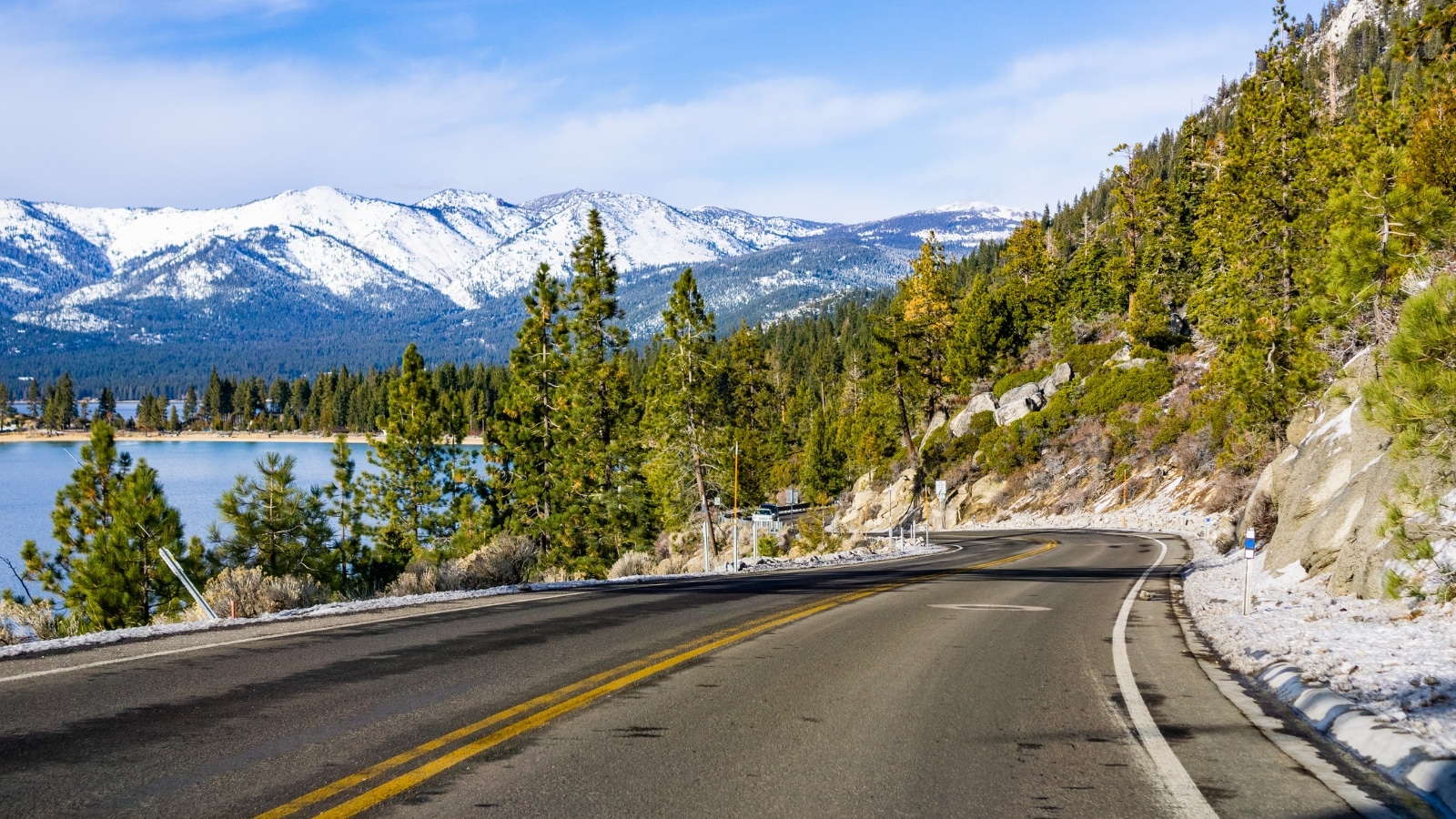 Lake Tahoe, showcasing the shoreline and Sierra mountains blanketed in snow. The scene features a winding road passing beneath the snow-capped mountains, flanked by greenery. Lake Tahoe is visible to the left of the picture.
