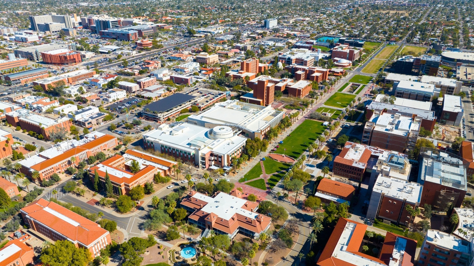 The aerial view of the University of Arizona's main campus in Tucson, Arizona, includes University Mall and the Old Main Building. The surrounding city of Tucson is visible, highlighting a mix of urban and residential areas.