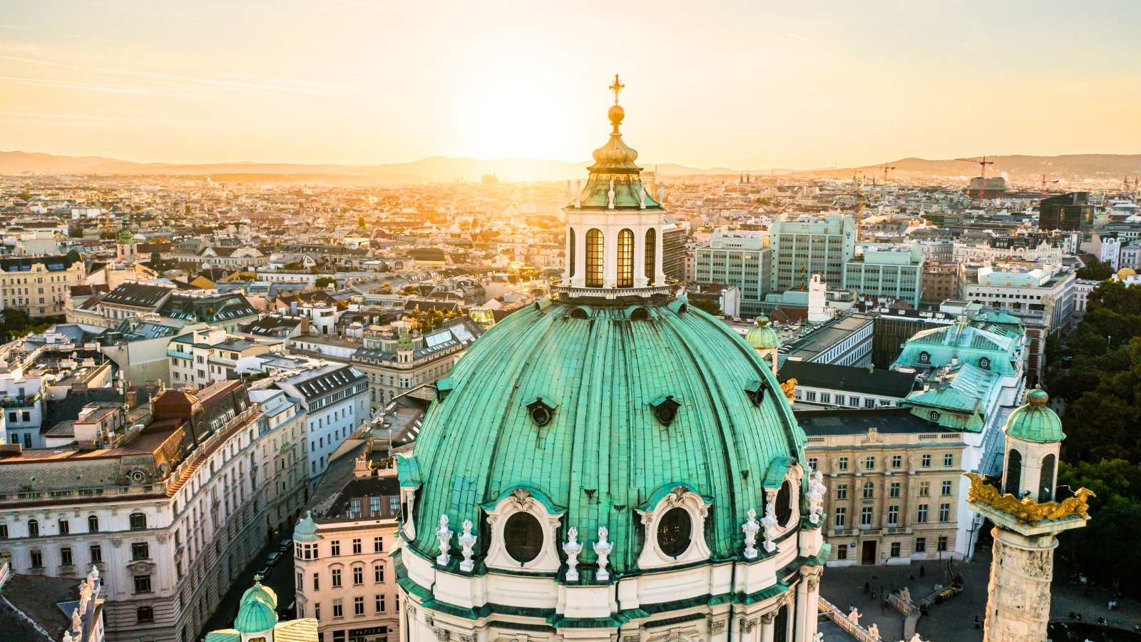 The aerial drone photo captures St. Charles Church "Karlskirche" in Vienna, Austria, during sunset. The church's magnificent Baroque architecture is highlighted against the evening sky. St. Charles Church features a large dome and intricate details on its façade, typical of Baroque design. In the background, a vast cityscape unfolds, showcasing Vienna's urban sprawl and architectural diversity.