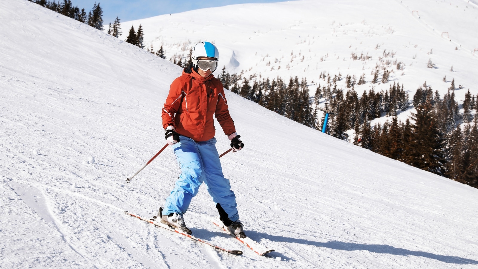 The image depicts a woman skiing downhill at a snowy resort. She is dressed in blue pants, a red jacket, and a white coat with a blue stripe. The surroundings showcase a typical winter scene with snow-covered slopes and trees.