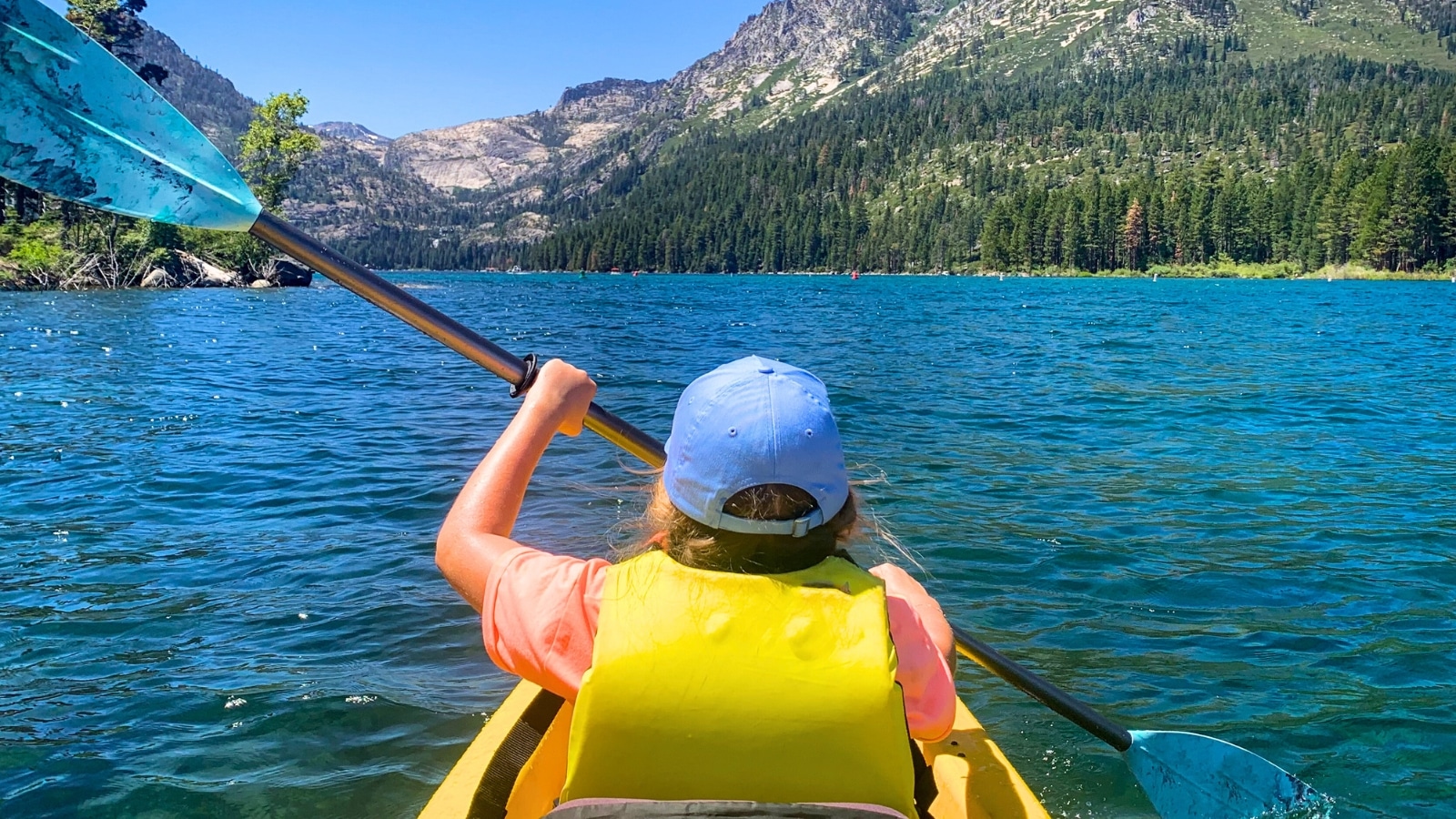 Young girl kayaking into Emerald Bay in Lake Tahoe, California. She is wearing a yellow water vest and a blue cap, holding a paddle. In the background, there are mountains typical of the Lake Tahoe region.