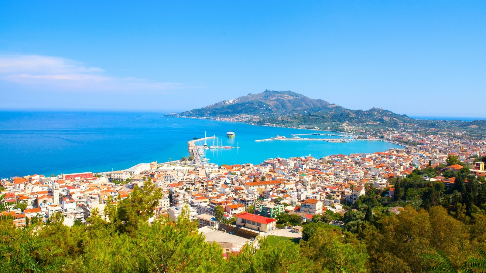 Port Zante in Zakynthos, Greece, offers a picturesque view of a cityscape with white houses and orange roofs in the foreground. The city overlooks a calm sea with a port area, characterized by a few boats and vessels. Hills rise in the background, adding a scenic backdrop to the coastal scene. The color scheme features the contrast of white buildings against the blue sea.