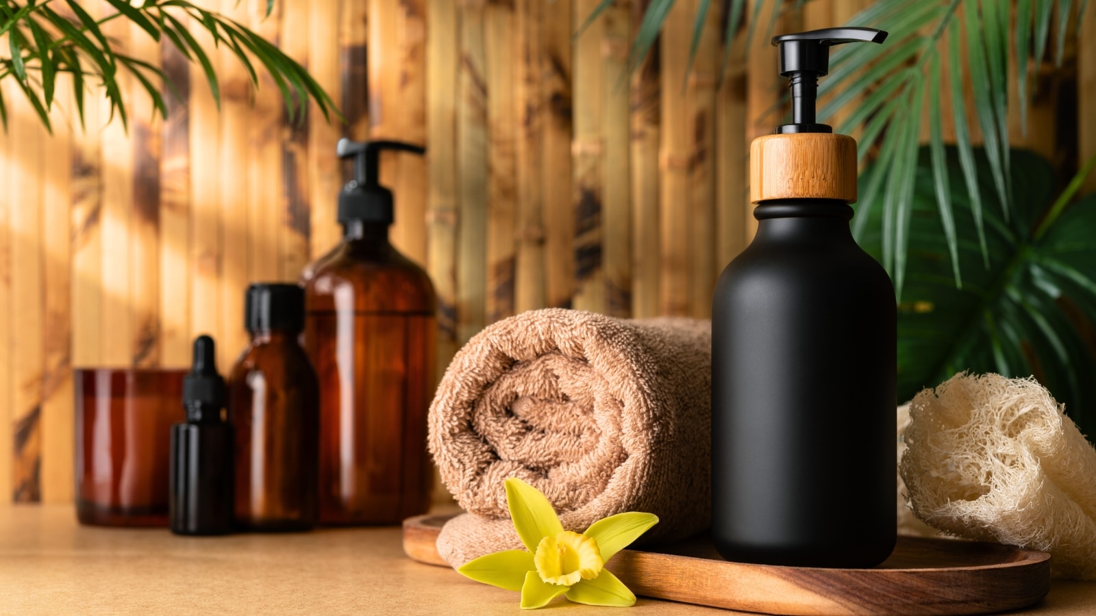 The image depicts a body products stand positioned against a bamboo wall, showcasing a variety of skincare and hygiene items. A rolled towel is placed nearby.