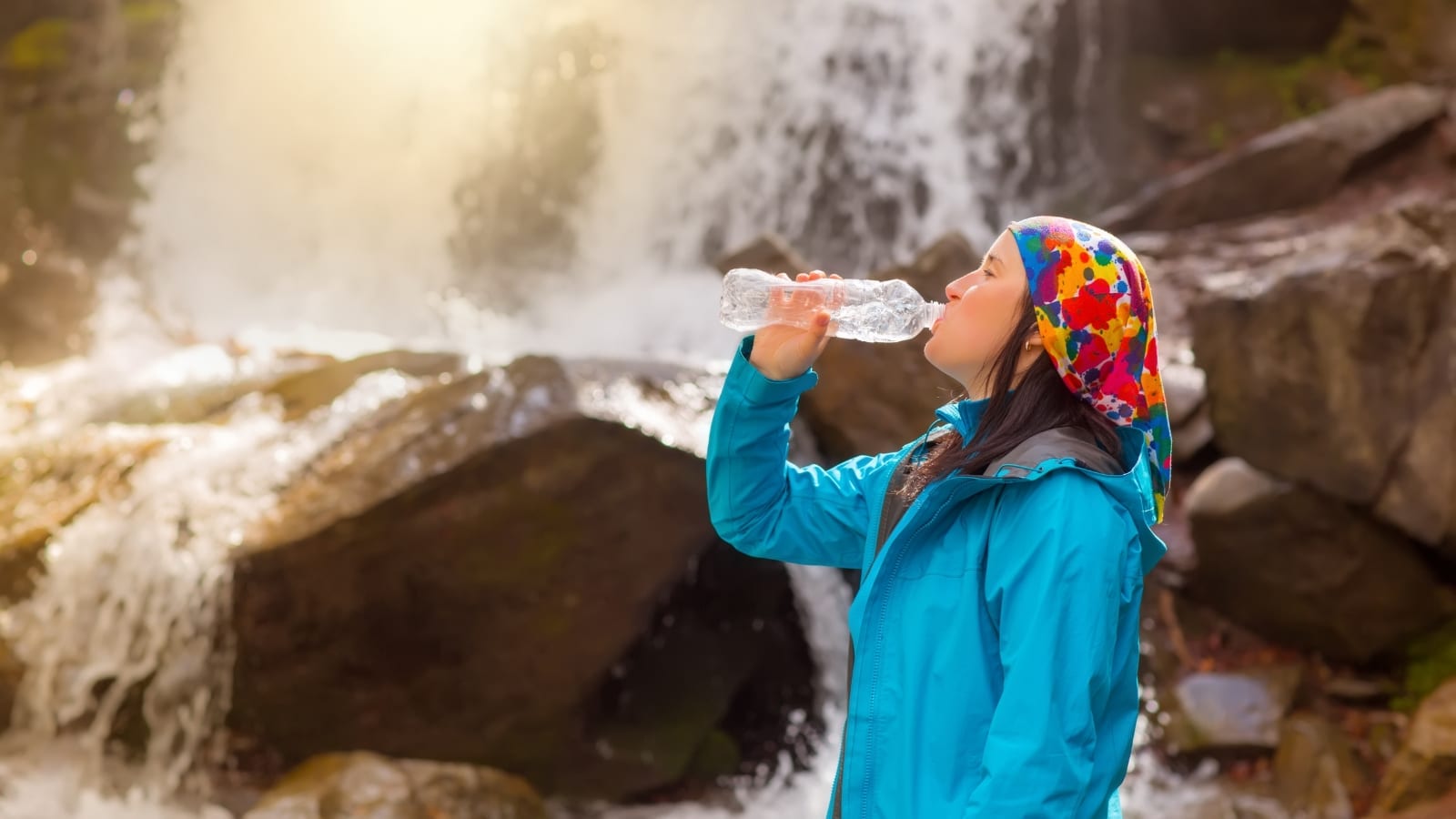 a woman is depicted against the backdrop of a waterfall, dressed in a blue hen with a colored scarf on her head. She drinking water from a transparent plastic bottle. 