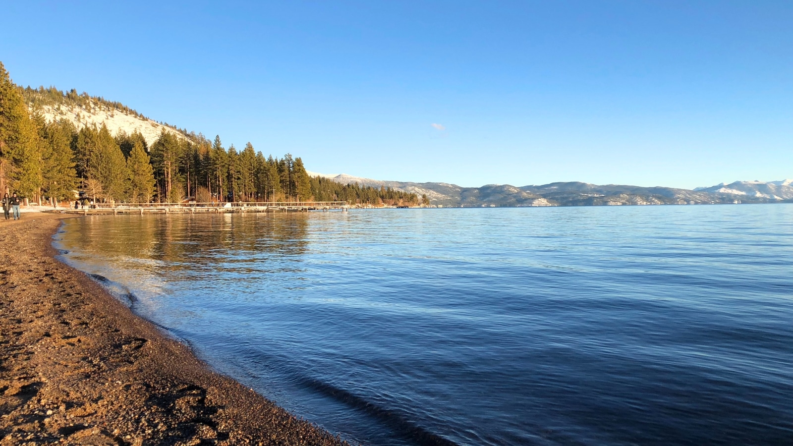 Kings Beach in North Lake Tahoe, featuring a clear distinction between the beach on the left and the pristine blue waters of the lake on the right. The water appears pure blue. In the background, hills rise, adorned with pine trees and additional foliage.