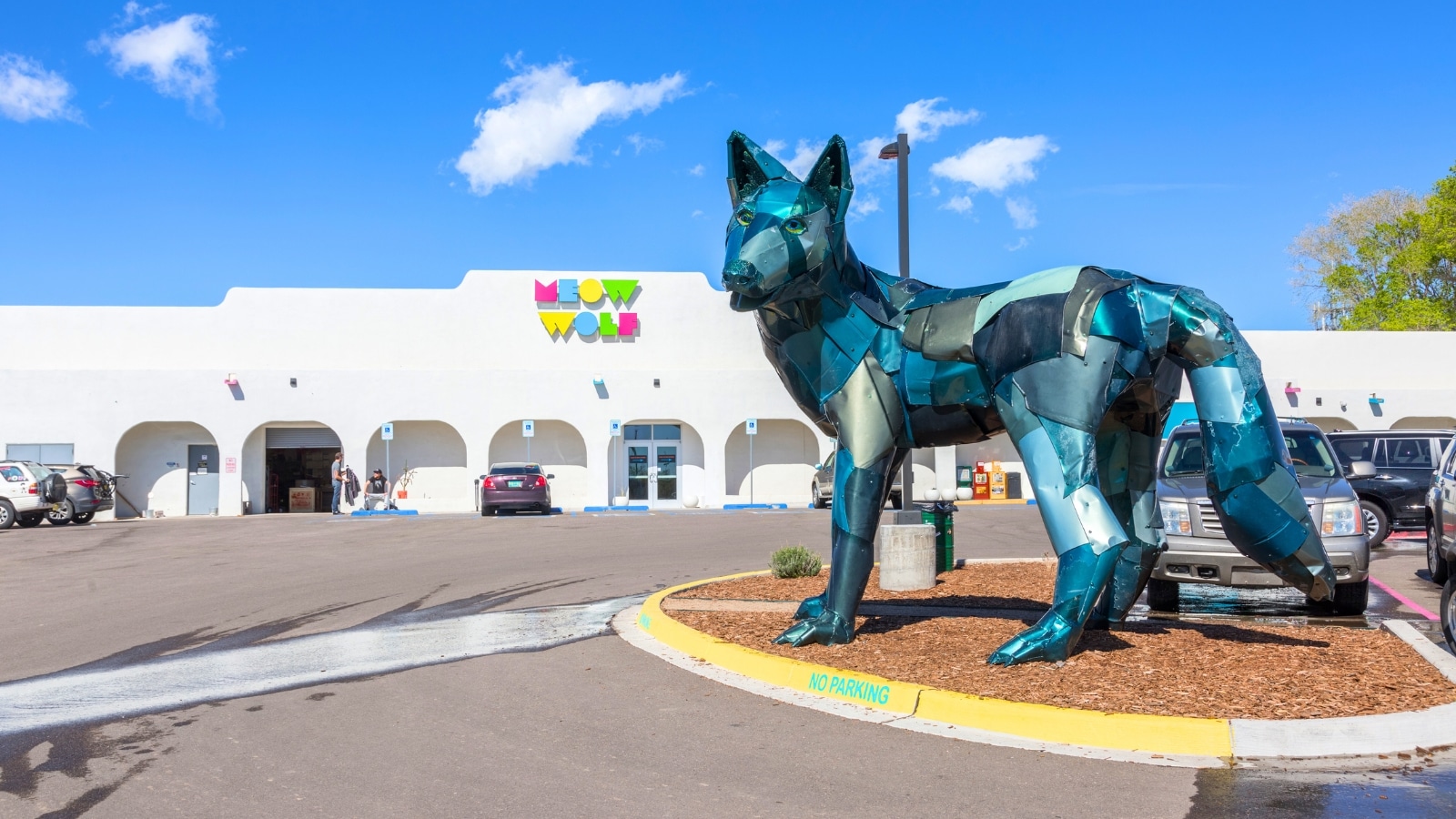 In Santa Fe, New Mexico, the Meow Wolf art collective features a blue wolf sculpture in the foreground. The background includes the Meow Wolf building, which is predominantly white with the name "Meow Wolf" displayed in colorful letters.