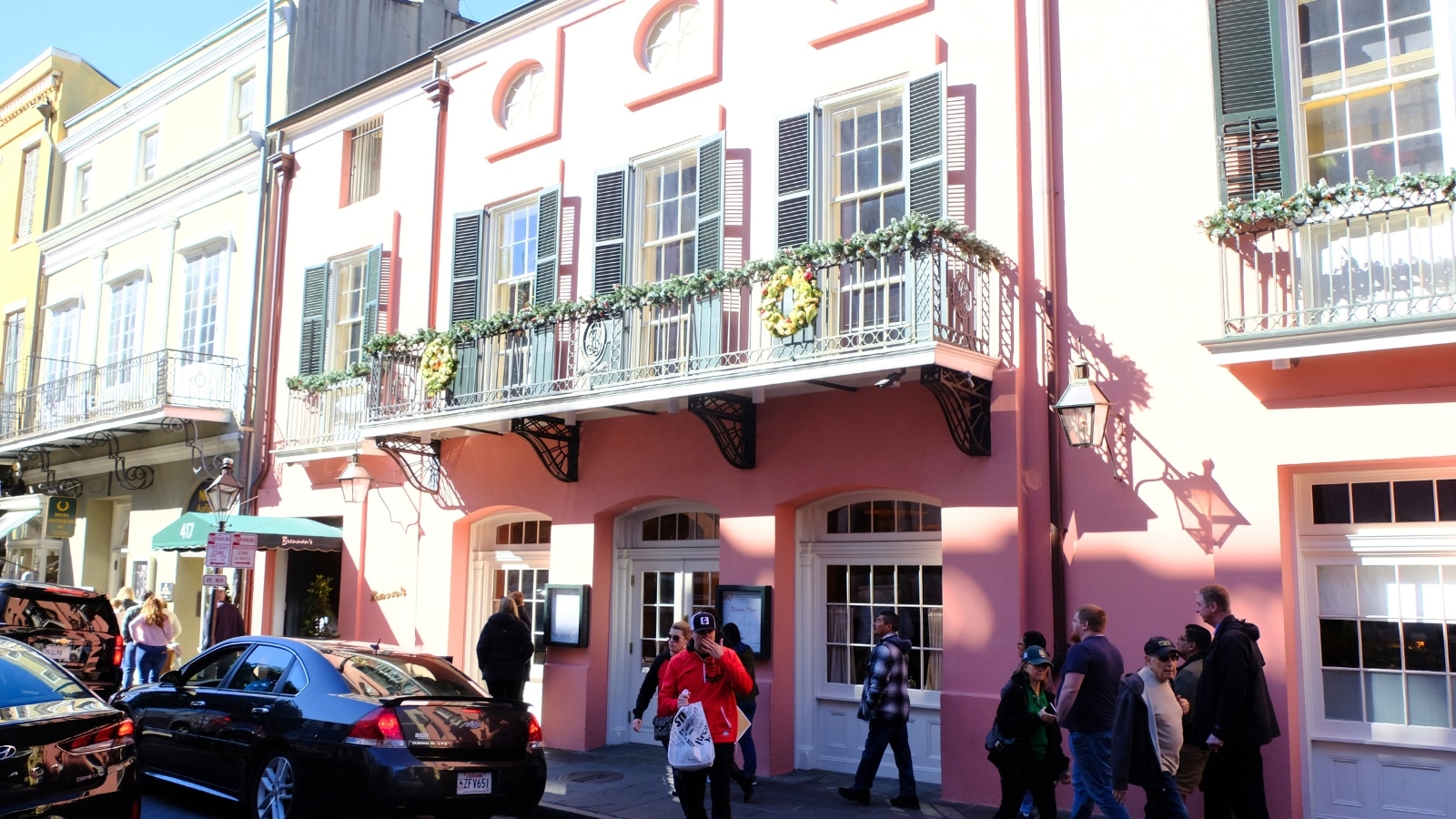 Brennan's Restaurant in New Orleans features a pink building with white shutters and doors. The street scene is bustling, with people walking by and cars driving past.