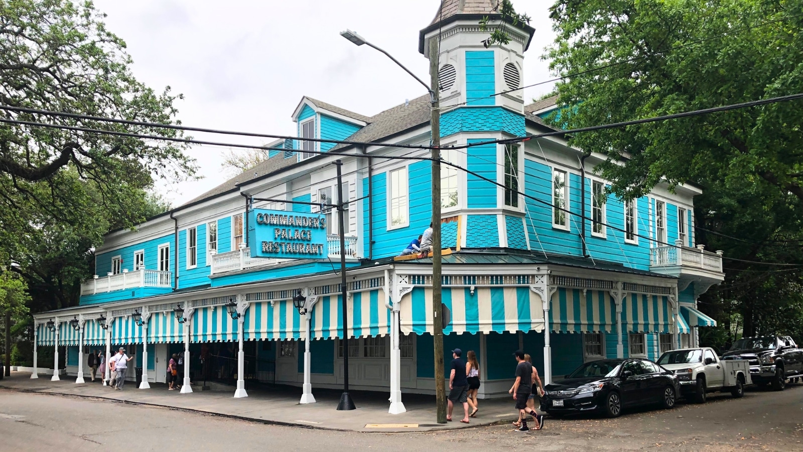 Commander's Palace in the Garden District of New Orleans features a turquoise building with white window frames. The street is lined with several parked cars, and people can be seen going about their activities. The sky appears overcast, adding a subdued atmosphere to the scene.