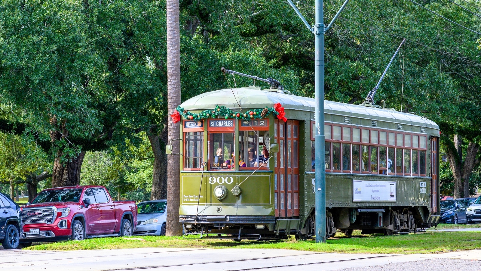 In New Orleans, USA, a tram travels along St. Charles Avenue, characterized by lush greenery including trees. The tram itself is dark green, blending with the surrounding foliage.