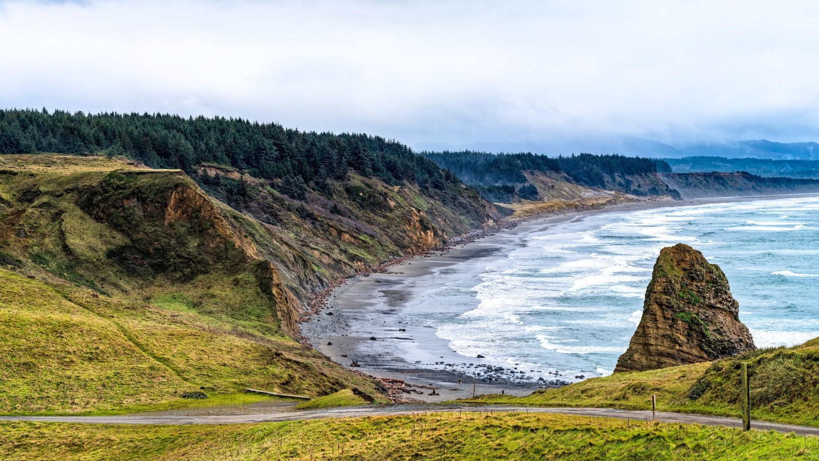 Cape Blanco State Park Beach in Oregon, USA. A winding road is depicted passing by a sea stack on the beach. The beach itself is long but not particularly wide, flanked by small hills and forested areas.