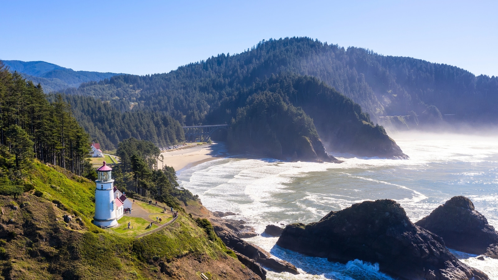 Heceta Head Light, a lighthouse located on the Oregon Coast. The lighthouse structure is prominently featured, standing tall against the coastal backdrop. The surrounding area includes a wide stretch of Florence Beach, flanked by numerous hills covered in lush vegetation.