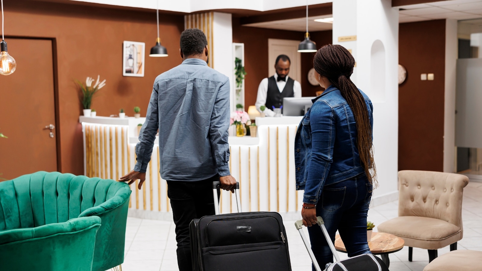 In the image, a young married couple is seen arriving at a resort hotel. They are depicted from the rear, pulling their luggage as they walk towards the front desk to check in.