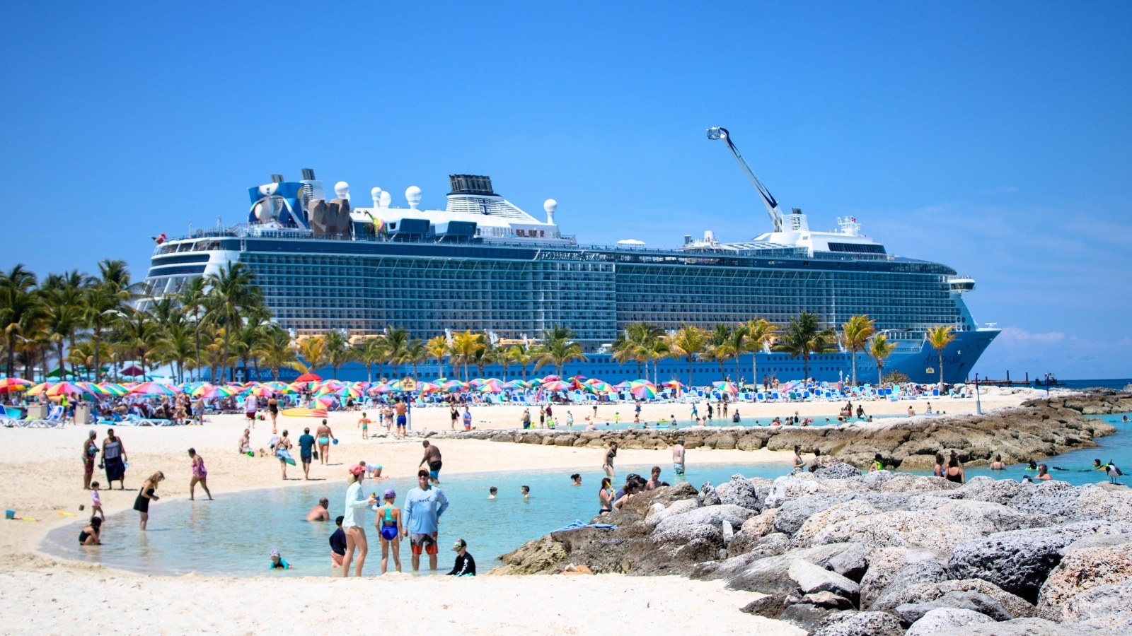 Bahamas with a Royal Caribbean International cruise ship in the background. In the foreground, a beach is visible, crowded with people and dotted with brightly colored umbrellas.