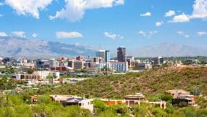 The image captures an aerial view of Tucson, Arizona, showcasing a beautiful panoramic scene under a clear sky with small, white clouds. In the background, hills add depth to the landscape.