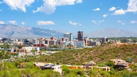 The image captures an aerial view of Tucson, Arizona, showcasing a beautiful panoramic scene under a clear sky with small, white clouds. In the background, hills add depth to the landscape.