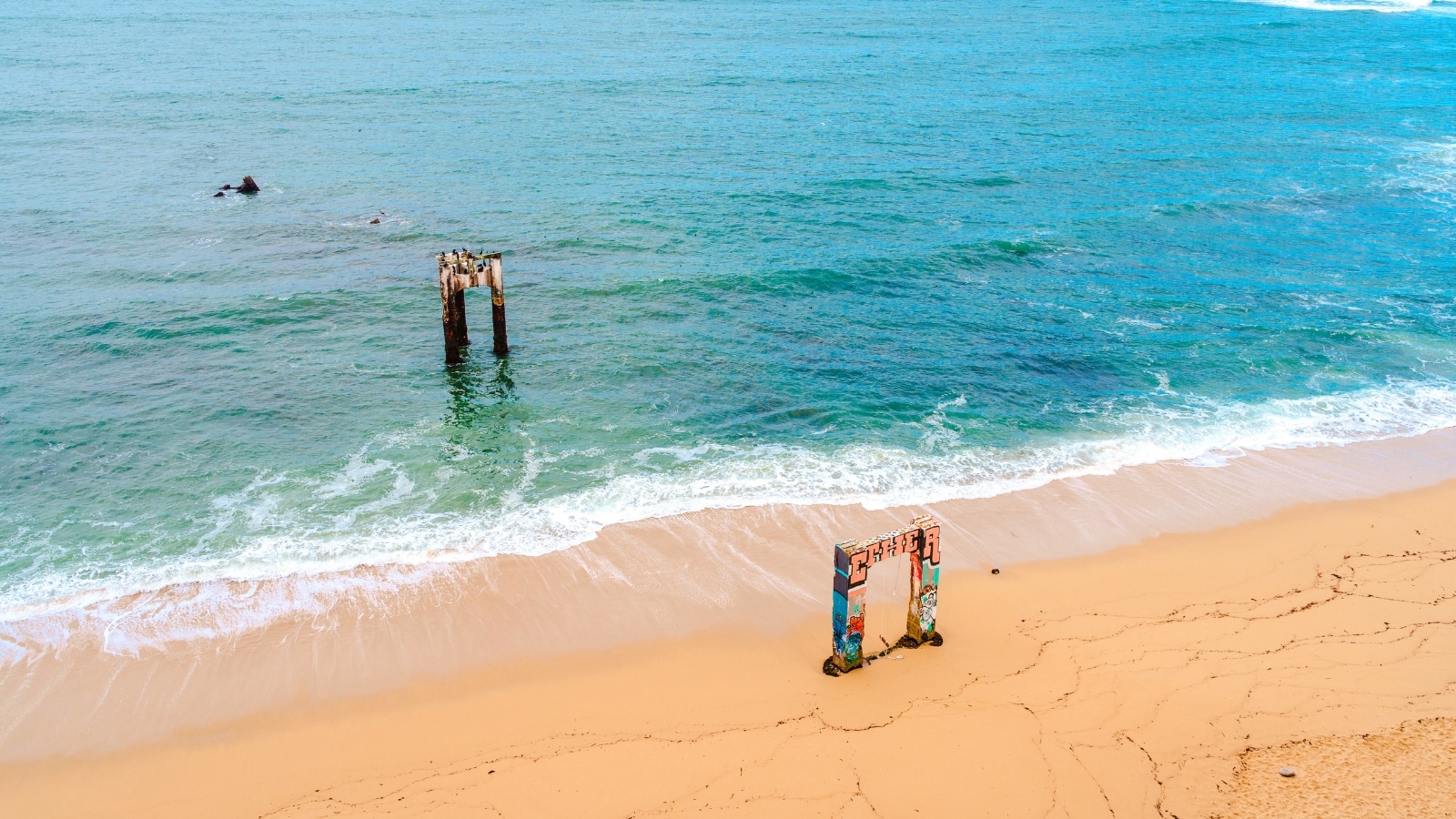The destroyed beams of a pier extend into the ocean in Davenport, California. The remnants of the pier are weathered and worn, with a rustic and abandoned appearance. The beams are a mix of dark, aged wood and corroded metal, jutting out of the water.