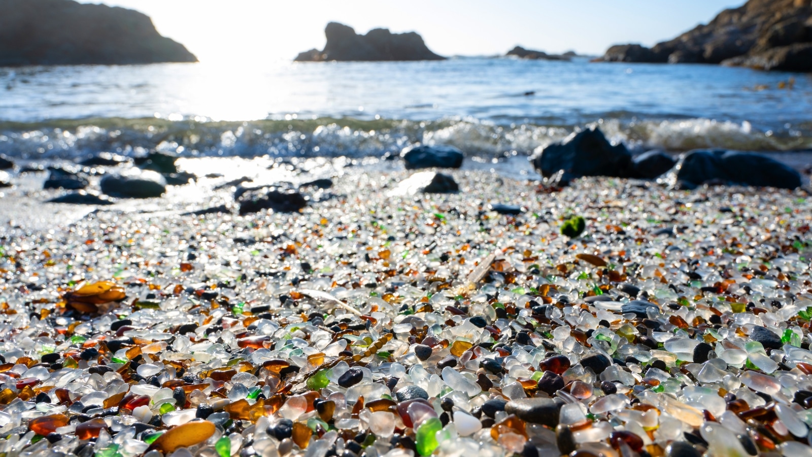 Millions of small pieces of old round-shaped glass cover the beach at Fort Bragg, California. The glass pieces create a colorful mosaic of greens, blues, whites, and browns. Ocean waves crash against the shore, with rocks scattered in the background.