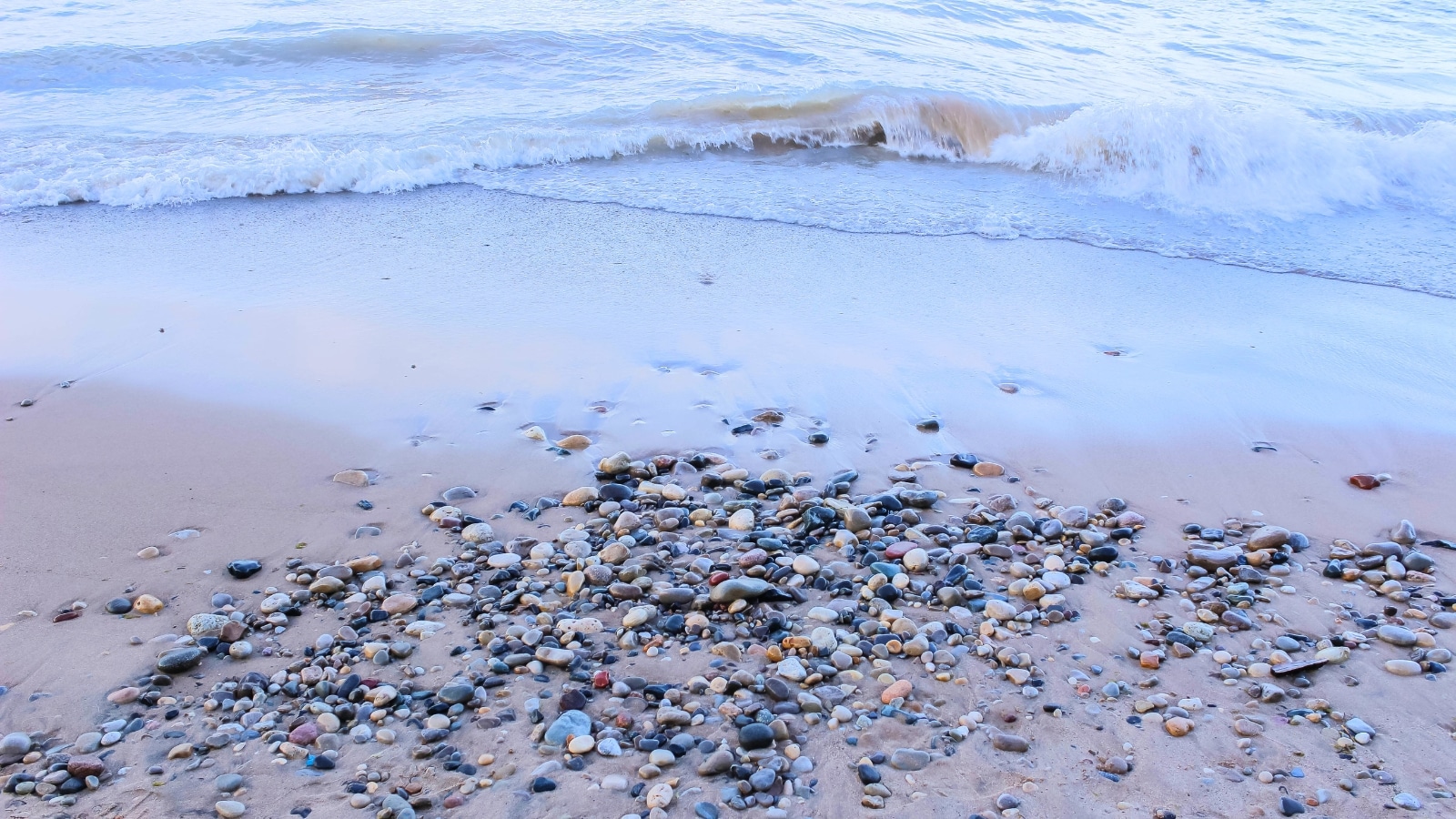 Waves crash onto the pebbled shoreline of Lake Michigan at Grant Park in Milwaukee, WI. The water is a deep blue, contrasting with the light-colored pebbles on the shore. The shoreline is lined with smooth, rounded stones of various sizes and colors.