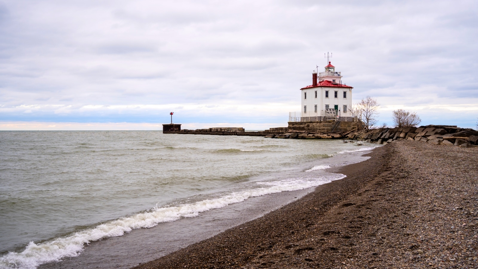 Fairport Harbor Lighthouse stands tall near the beach, under a cloudy sky. The lighthouse features a classic white tower with a red roof, contrasting against the grey clouds. Surrounding the lighthouse, the beach is sandy with scattered rocks and pebbles.