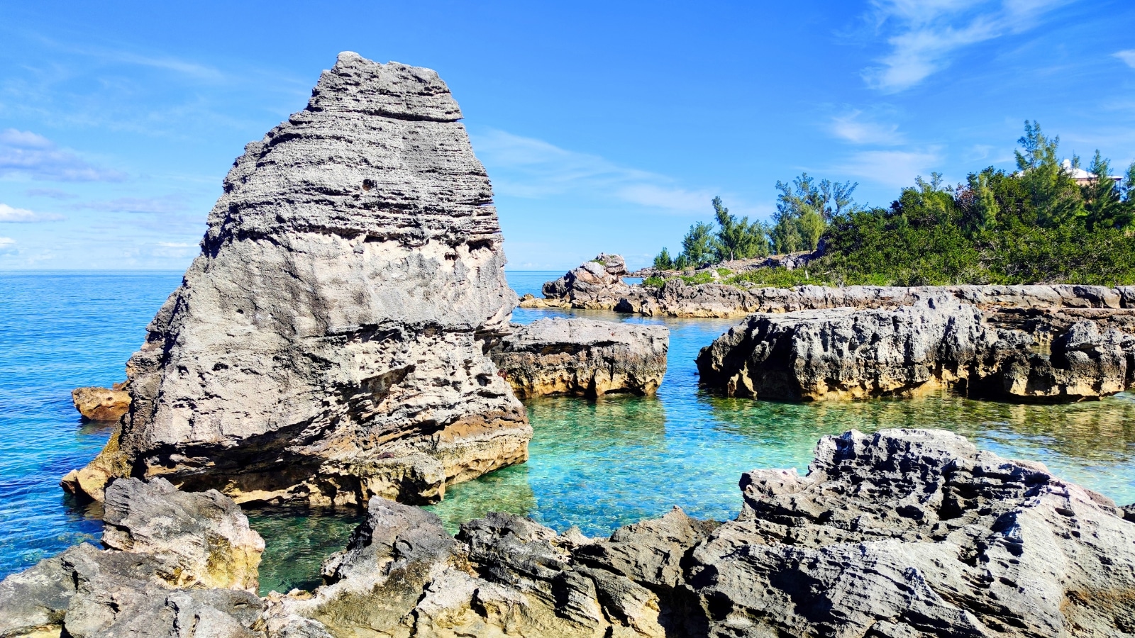 Bermuda Island features a tropical coastal landscape with vibrant turquoise waters and white sandy beaches. The coastline is dotted with lush green vegetation, including trees and flowering shrubs. Rocky outcrops and coral reefs are visible near the shore, adding texture and color to the scene.
